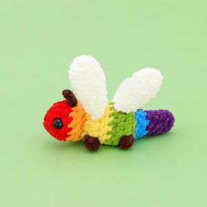 May include: A small, crocheted insect toy featuring a rainbow-colored body with segments in red, orange, yellow, green, blue, and purple. It has two white, textured wings, two small dark brown antennae, four tiny dark brown legs, and a black eye.