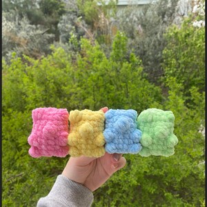 May include: Four crocheted stuffed toys in pink, yellow, blue, and green. The toys are in a row and are being held by a hand.