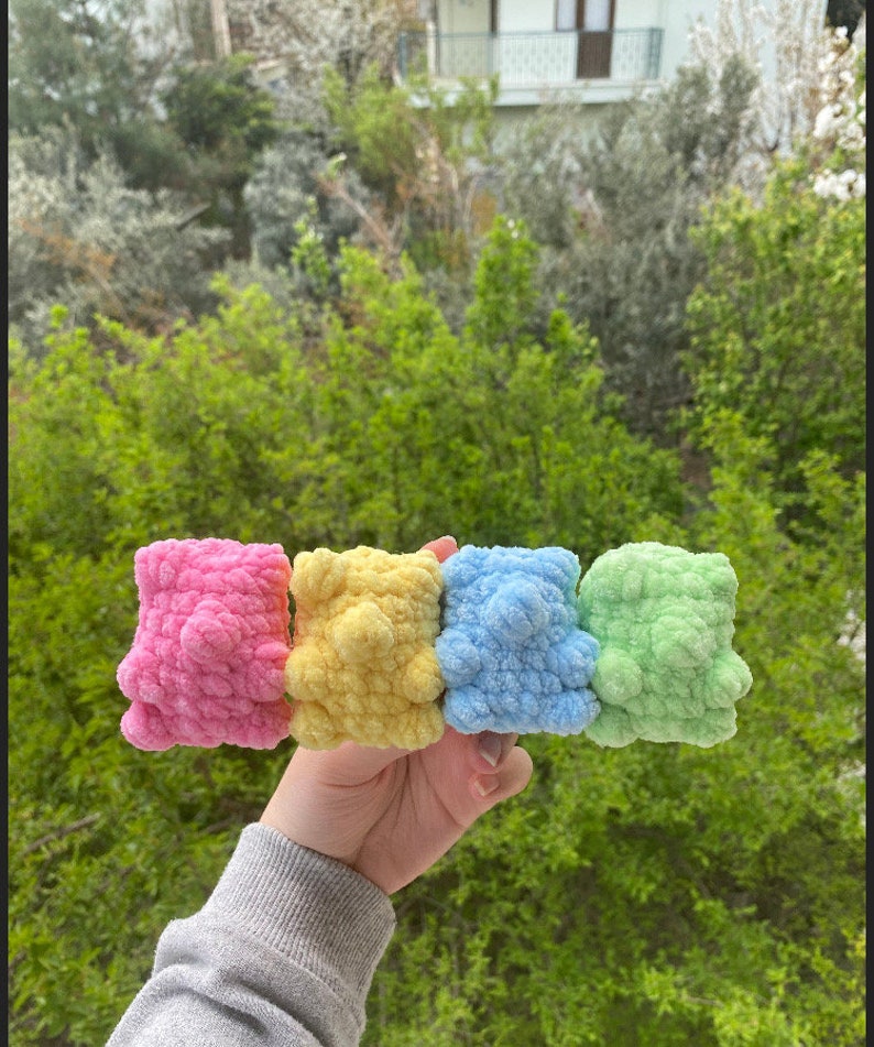 May include: Four crocheted stuffed toys in pink, yellow, blue, and green. The toys are in a row and are being held by a hand.