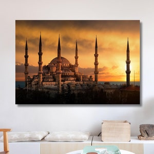 May include: A canvas print of the Blue Mosque in Istanbul, Turkey, at sunset. The image features the mosque's minarets and domes silhouetted against a vibrant orange and yellow sky. The artwork is displayed in a modern living room setting.