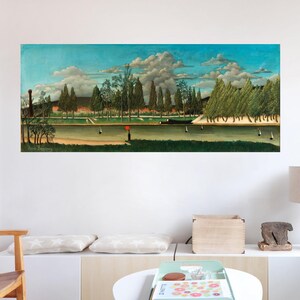 May include: A painting of a landscape with a river, trees, and a blue sky with white clouds. The painting is framed and hanging on a white wall in a living room.