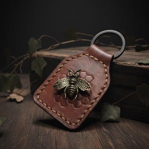 May include: A brown leather keychain with a metal bee emblem. The keychain has a stitched border and a metal ring. The bee is detailed with wings and body segments. The background is a dark wooden surface.