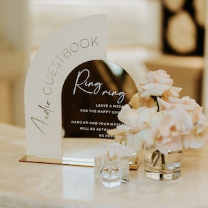 Op de afbeelding: Een gouden spiegel met de tekst "Ring ring" en "Leave a message for the happy couple" erop. De spiegel staat op een witte tafel met een kleine vaas met roze rozen en een glazen doos met een ring erin.