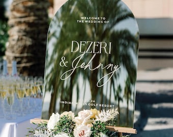 May include: An arched, reflective wedding welcome sign on a wooden easel, featuring the text "WELCOME TO THE WEDDING OF DEZERI & Johnny" and "04.13.24 INDIAN WELLS GOLF RESORT," adorned with a large arrangement of white and cream roses and greenery.