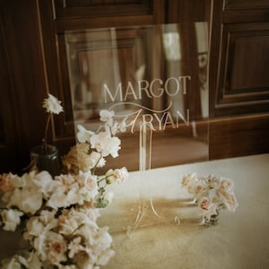 K&ouml;nnte beinhalten: Eine transparente Acryltafel mit den Namen "MARGOT" und "RYAN" in eleganter wei&szlig;er Schrift. Das Schild steht auf einem transparenten St&auml;nder, umgeben von zarten rosa und wei&szlig;en Blumen, was eine zarte und romantische &Auml;sthetik erzeugt.