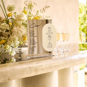 May include: A bar scene with a floral arrangement of white and yellow flowers, a menu with the word "Ciao!", and three champagne flutes filled with a golden liquid. The menu lists beer, wine, and spirits.