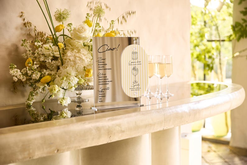 May include: A bar scene with a floral arrangement of white and yellow flowers, a menu with the word "Ciao!", and three champagne flutes filled with a golden liquid. The menu lists beer, wine, and spirits.