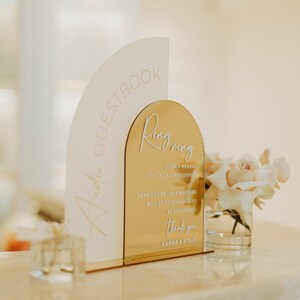 Op de afbeelding: Een gouden en wit bord met de tekst "Guestbook" en "Ring Ring" erop. Het bord bevat ook de tekst "Laat een bericht achter voor het gelukkige paar" en "Bedankt Aaron & Kylie".