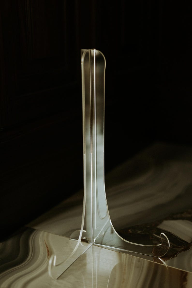 K&ouml;nnte beinhalten: Ein klarer Acryl-Displayst&auml;nder mit einem hohen, schlanken Design. Der St&auml;nder hat eine gebogene Basis und eine vertikale St&uuml;tzstruktur mit drei parallelen Linien. Der dunkle Hintergrund hebt die transparente Form hervor.