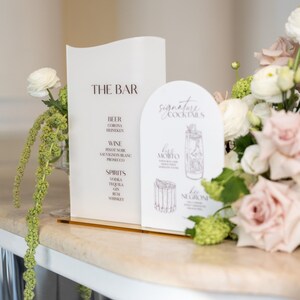 May include: A white bar menu and a signature cocktail menu with a gold base. The bar menu lists beer, wine, and spirits. The cocktail menu features a Mojito and Negroni illustration. Floral arrangements are in the background.