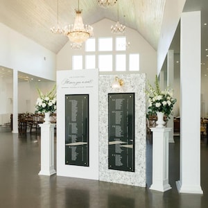 Puede incluir: Dos pilares blancos flanquean un plano de asientos de boda. La tabla es negra con texto blanco, y el fondo es blanco con un estampado floral. El texto dice "Since you a seat". Candelabros cuelgan del techo alto.