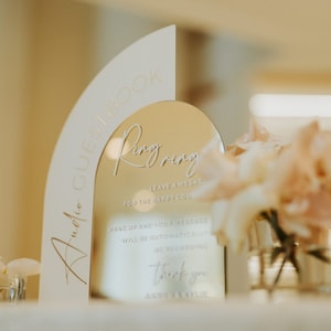 Op de afbeelding: Een gouden spiegel met een wit frame in de vorm van een boog. De spiegel heeft de tekst "Ring Ring" erop, samen met instructies voor gasten om een ​​bericht achter te laten voor het gelukkige paar. De tekst "Audio Guestbook" staat op het witte frame.