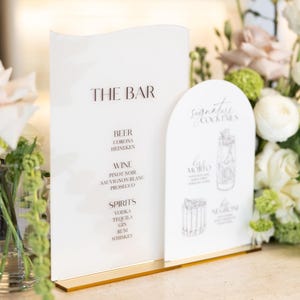 May include: A white bar menu with gold accents lists drinks: beer, wine, and spirits. A separate card features signature cocktails with illustrations. The text "THE BAR" is at the top of the menu.
