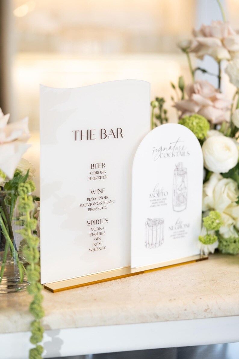 May include: A white bar menu with gold accents lists drinks: beer, wine, and spirits. A separate card features signature cocktails with illustrations. The text "THE BAR" is at the top of the menu.