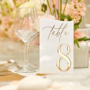 May include: White table number card with gold lettering that says "Table" and the number "8".