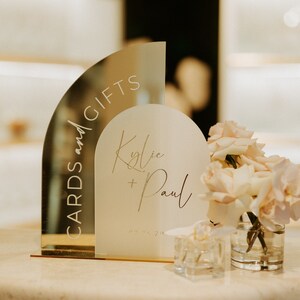 May include: A gold and white acrylic sign with the text "Cards and Gifts" and "Kylie + Paul" in a modern calligraphy font. The sign is decorated with a bouquet of pale pink roses and orchids in a clear glass vase.