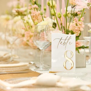 May include: A white table number card with gold calligraphy that reads "Table 8" on a table setting with white flowers and clear glassware.