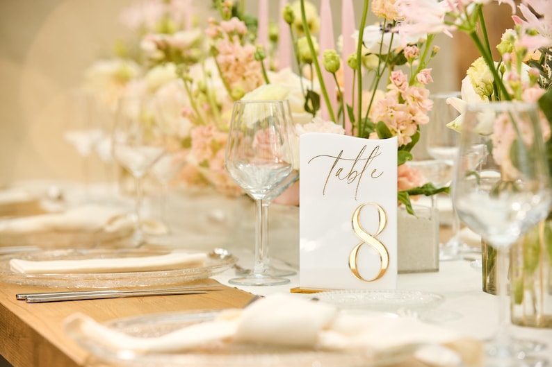 May include: A white table number card with gold calligraphy that reads "Table 8" on a table setting with white flowers and clear glassware.