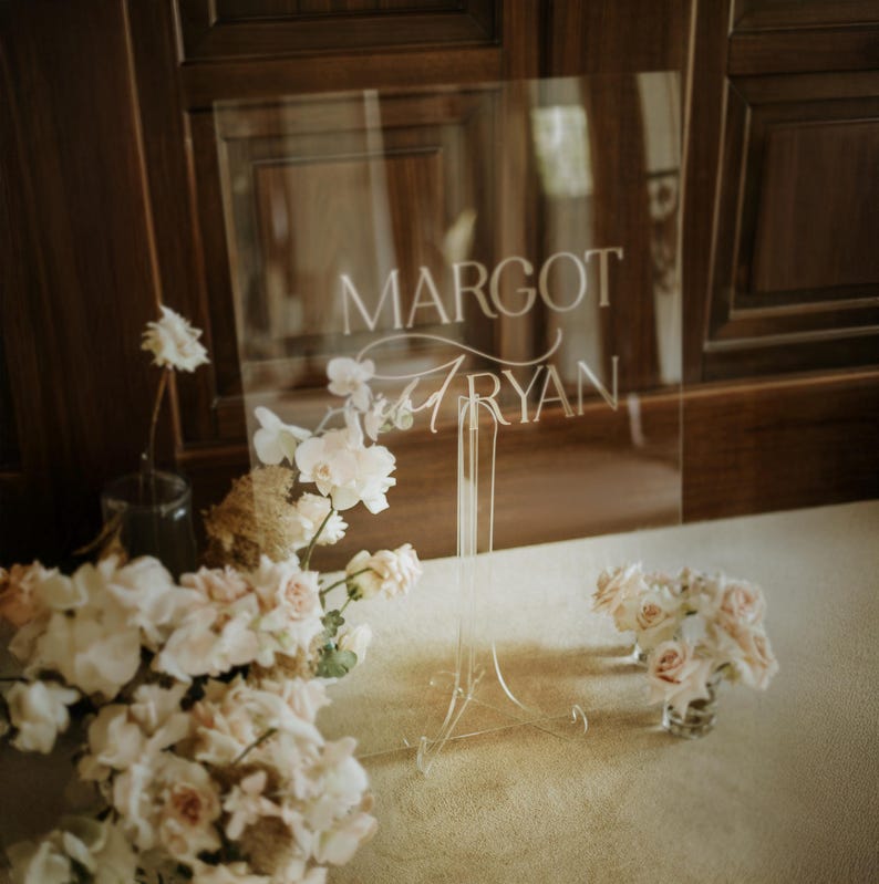 K&ouml;nnte beinhalten: Eine transparente Acryltafel mit den Namen "MARGOT" und "RYAN" in eleganter wei&szlig;er Schrift. Das Schild steht auf einem transparenten St&auml;nder, umgeben von zarten rosa und wei&szlig;en Blumen, was eine zarte und romantische &Auml;sthetik erzeugt.