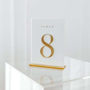 Può includere: Un segnaposto bianco con la scritta «TABLE» in alto e il numero «8» in oro. Il segnaposto è su un supporto in acrilico trasparente con base dorata. Lo sfondo è bianco.