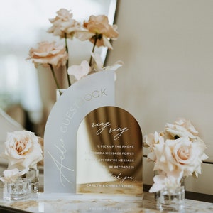 Op de afbeelding: Een gouden en wit acryl bord met de tekst "Audio Guestbook" en "ring ring" met instructies om een bericht voor het paar op te nemen. Het bord is versierd met roze rozen.
