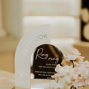 Op de afbeelding: Een gouden acryl bord met een gespiegelde boog. Het bord heeft de tekst "Ring Ring" en bevat instructies voor gasten om een bericht achter te laten voor het gelukkige paar. Het bord staat op een wit oppervlak met een vaas met roze rozen.