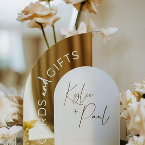 May include: Gold and white acrylic wedding sign with the text "Cards and Gifts" on the gold side and "Kylie + Paul 07.24.2023" on the white side.