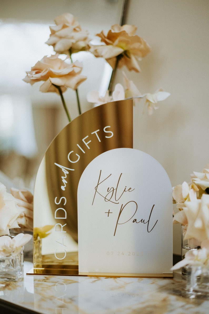 May include: Gold and white acrylic wedding sign with the text "Cards and Gifts" on the gold side and "Kylie + Paul 07.24.2023" on the white side.