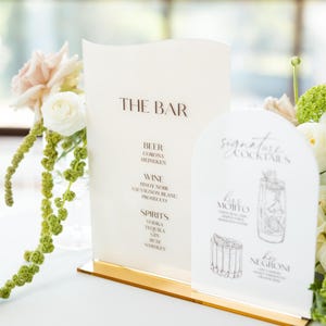 May include: A bar menu with the text "THE BAR" in brown lettering. The menu lists beer, wine, and spirits. A smaller sign displays signature cocktails, including a Mojito and a Negroni. The signs are on a gold base.