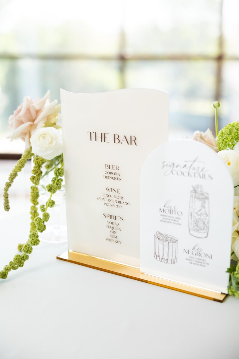 May include: A bar menu with the text "THE BAR" in brown lettering. The menu lists beer, wine, and spirits. A smaller sign displays signature cocktails, including a Mojito and a Negroni. The signs are on a gold base.