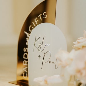 May include: Gold and white acrylic wedding sign with the text "Cards and Gifts" and "Kylie + Paul".