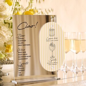 May include: A clear acrylic bar menu with the word "Ciao!" at the top. The menu lists beer, wine, and spirits. A smaller, striped sign advertises a "Lemon love" cocktail and a "Spritz" cocktail. Champagne flutes are in the background.