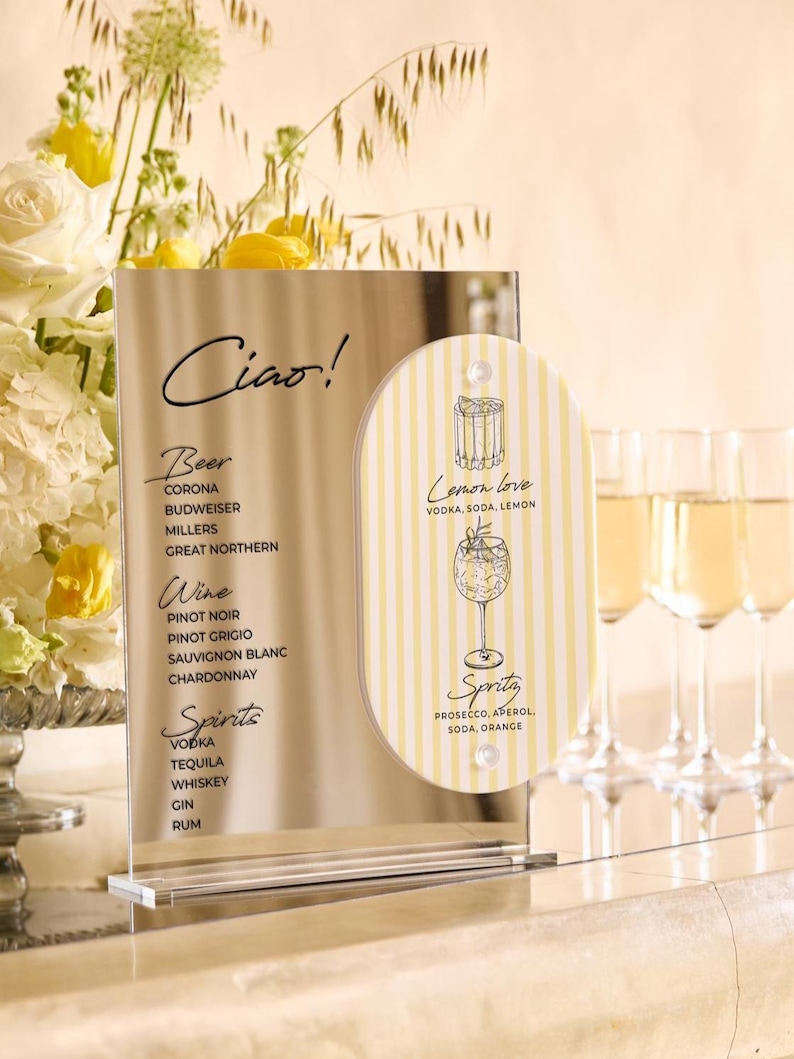 May include: A clear acrylic bar menu with the word "Ciao!" at the top. The menu lists beer, wine, and spirits. A smaller, striped sign advertises a "Lemon love" cocktail and a "Spritz" cocktail. Champagne flutes are in the background.