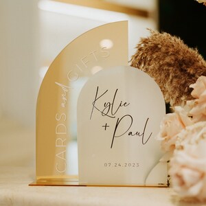 May include: A white acrylic sign with the text "Kylie + Paul" and the date "07.24.2023" in black script. A gold acrylic sign with the text "Cards and Gifts" is behind the white sign.