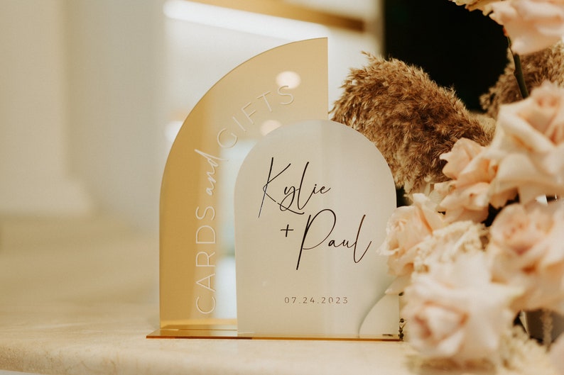 May include: A white acrylic sign with the text "Kylie + Paul" and the date "07.24.2023" in black script. A gold acrylic sign with the text "Cards and Gifts" is behind the white sign.