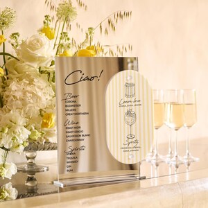 May include: A clear acrylic bar menu with the word "Ciao!" at the top. The menu lists beer, wine, and spirits. A smaller, striped sign says "Lemon love" and "Spritz" with a cocktail illustration. Champagne flutes are in the background.