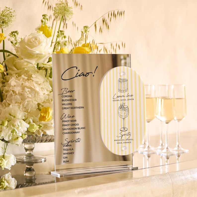 May include: A clear acrylic bar menu with the word "Ciao!" at the top. The menu lists beer, wine, and spirits. A smaller, striped sign says "Lemon love" and "Spritz" with a cocktail illustration. Champagne flutes are in the background.