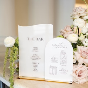 May include: Two white signs with gold bases display bar menus. One lists beer, wine, and spirits. The other features signature cocktails, including a Mojito and Negroni, with illustrations. Floral arrangements of pink and white roses are in the background.