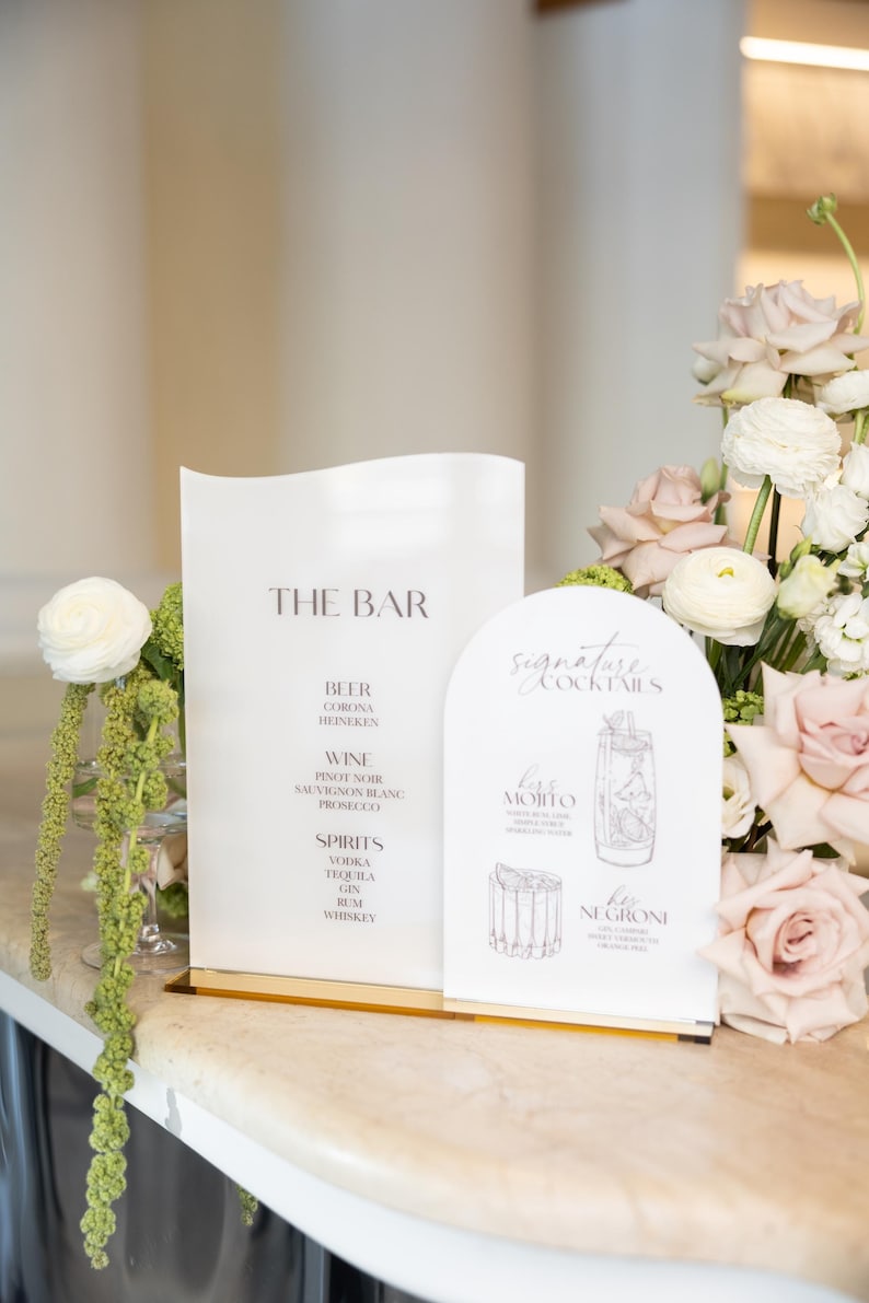 May include: Two white signs with gold bases display bar menus. One lists beer, wine, and spirits. The other features signature cocktails, including a Mojito and Negroni, with illustrations. Floral arrangements of pink and white roses are in the background.