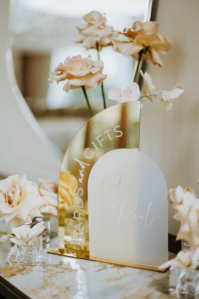 May include: A gold mirrored sign with the text "Cards and Gifts" and a white frosted sign with the text "Katie & Paul" in cursive. The signs are decorated with pale pink flowers.