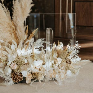 K&ouml;nnte beinhalten: Ein klarer Acrylschild mit einem Blumengesteck. Das Arrangement besteht aus getrocknetem Pampasgras, wei&szlig;en Rosen und anderen getrockneten Blumen in Creme- und Beiget&ouml;nen. Das Schild ist f&uuml;r Hochzeiten oder Veranstaltungen konzipiert.