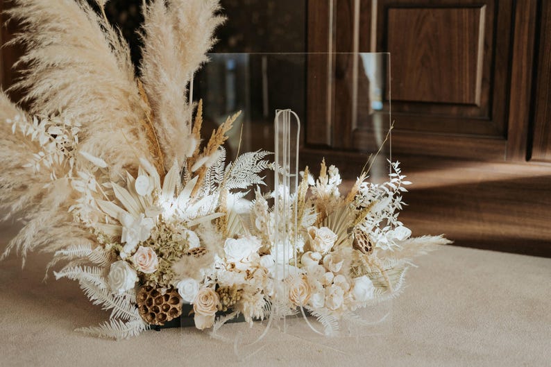 K&ouml;nnte beinhalten: Ein klarer Acrylschild mit einem Blumengesteck. Das Arrangement besteht aus getrocknetem Pampasgras, wei&szlig;en Rosen und anderen getrockneten Blumen in Creme- und Beiget&ouml;nen. Das Schild ist f&uuml;r Hochzeiten oder Veranstaltungen konzipiert.