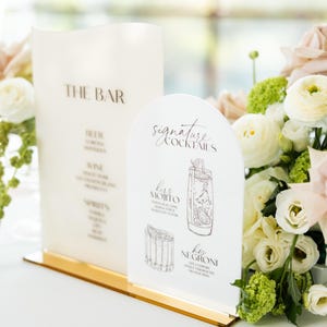 May include: A bar menu and signature cocktail signs with a gold base. The bar menu lists beer, wine, and spirits. The cocktail sign features illustrations of a Mojito and Negroni. Floral arrangements in the background.