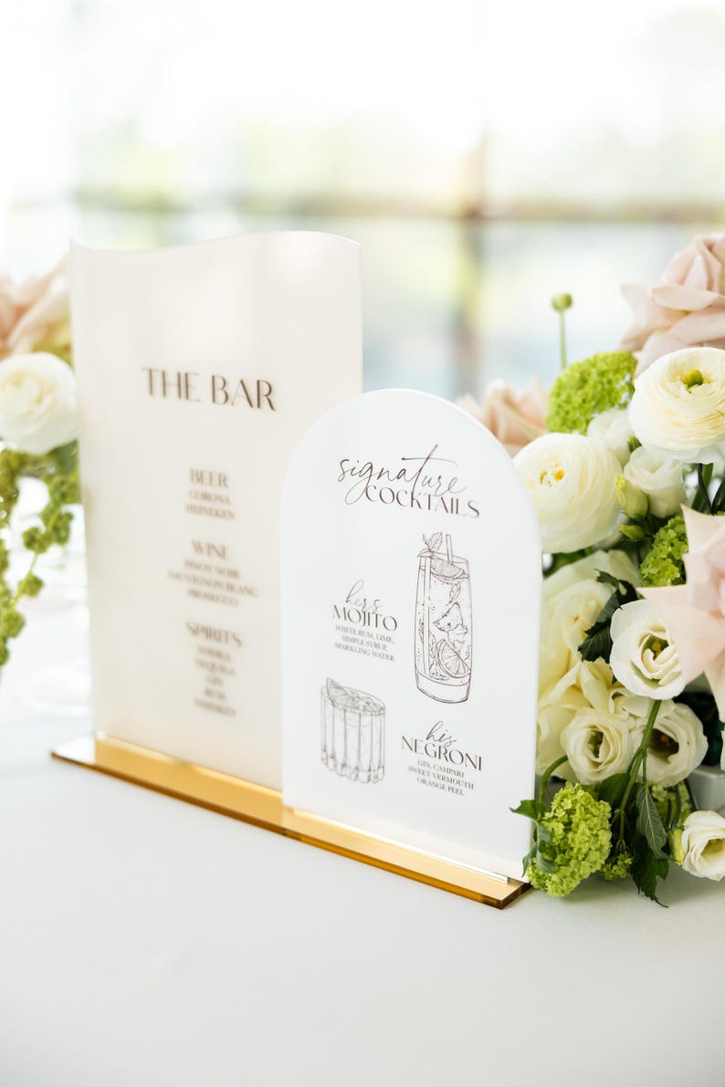 May include: A bar menu and signature cocktail signs with a gold base. The bar menu lists beer, wine, and spirits. The cocktail sign features illustrations of a Mojito and Negroni. Floral arrangements in the background.