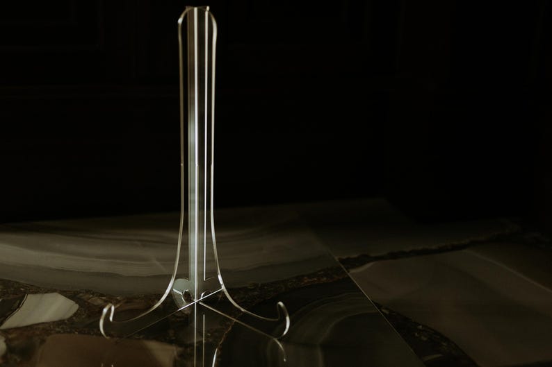 K&ouml;nnte beinhalten: Ein transparenter Acryl-Pr&auml;sentationsst&auml;nder. Der St&auml;nder hat ein hohes, schlankes Design mit einer Basis mit gebogenen St&uuml;tzen und einer vertikalen St&uuml;tze mit mehreren parallelen Linien. Der dunkle Hintergrund hebt die Form des St&auml;nders hervor.