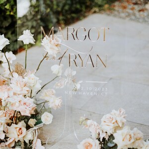 K&ouml;nnte beinhalten: Transparente Hochzeitsanzeige mit goldfarbenem Schriftzug &bdquo;Margot and Ryan&ldquo;, dem Datum 12.10.2023 und dem Ort New Haven, CT. Das Schild ist mit hellrosa und wei&szlig;en Rosen und anderen floralen Arrangements geschm&uuml;ckt.