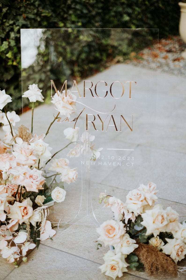 K&ouml;nnte beinhalten: Transparente Hochzeitsanzeige mit goldfarbenem Schriftzug &bdquo;Margot and Ryan&ldquo;, dem Datum 12.10.2023 und dem Ort New Haven, CT. Das Schild ist mit hellrosa und wei&szlig;en Rosen und anderen floralen Arrangements geschm&uuml;ckt.
