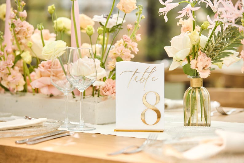 May include: A white table number card with gold lettering that reads "Table 8" is displayed on a table with a floral centrepiece. The centrepiece features pink and white flowers in a clear vase.
