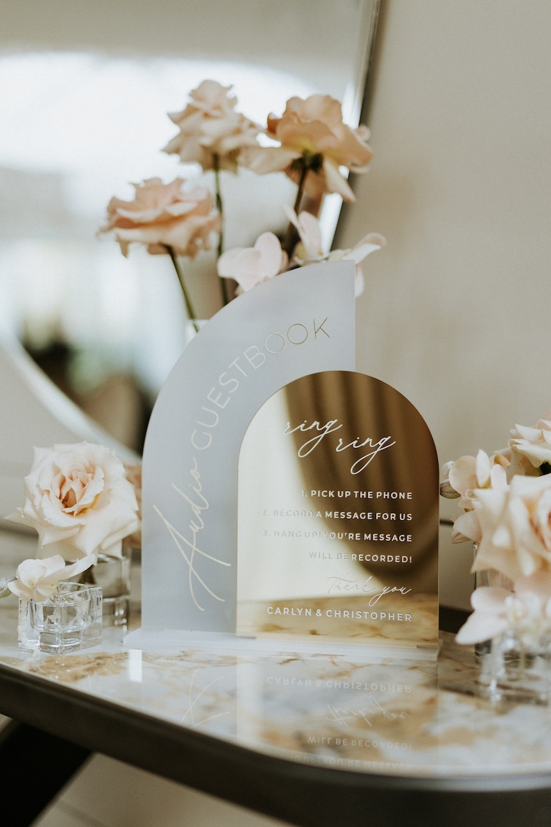 Op de afbeelding: Een gouden en wit acryl bord met de tekst "Audio Guestbook" en instructies voor het opnemen van een bericht. Het bord is boogvormig en heeft een spiegelend oppervlak. De tekst "Ring Ring" is in cursief geschreven op het spiegelende oppervlak. Het bord staat op een marmeren tafel met roze bloemen op de achtergrond.