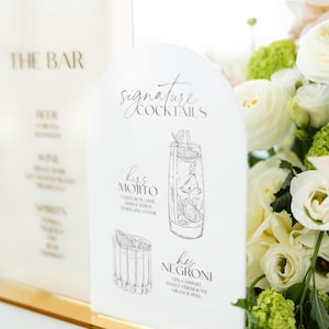 May include: White acrylic bar signs with gold trim. One sign reads "THE BAR" with a list of drinks. The other sign says "Signature Cocktails" and features illustrations of a Mojito and Negroni, with their ingredients.